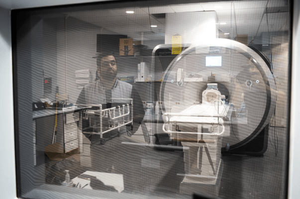 The reflection of a man is seen looking through a window at an MRI machine